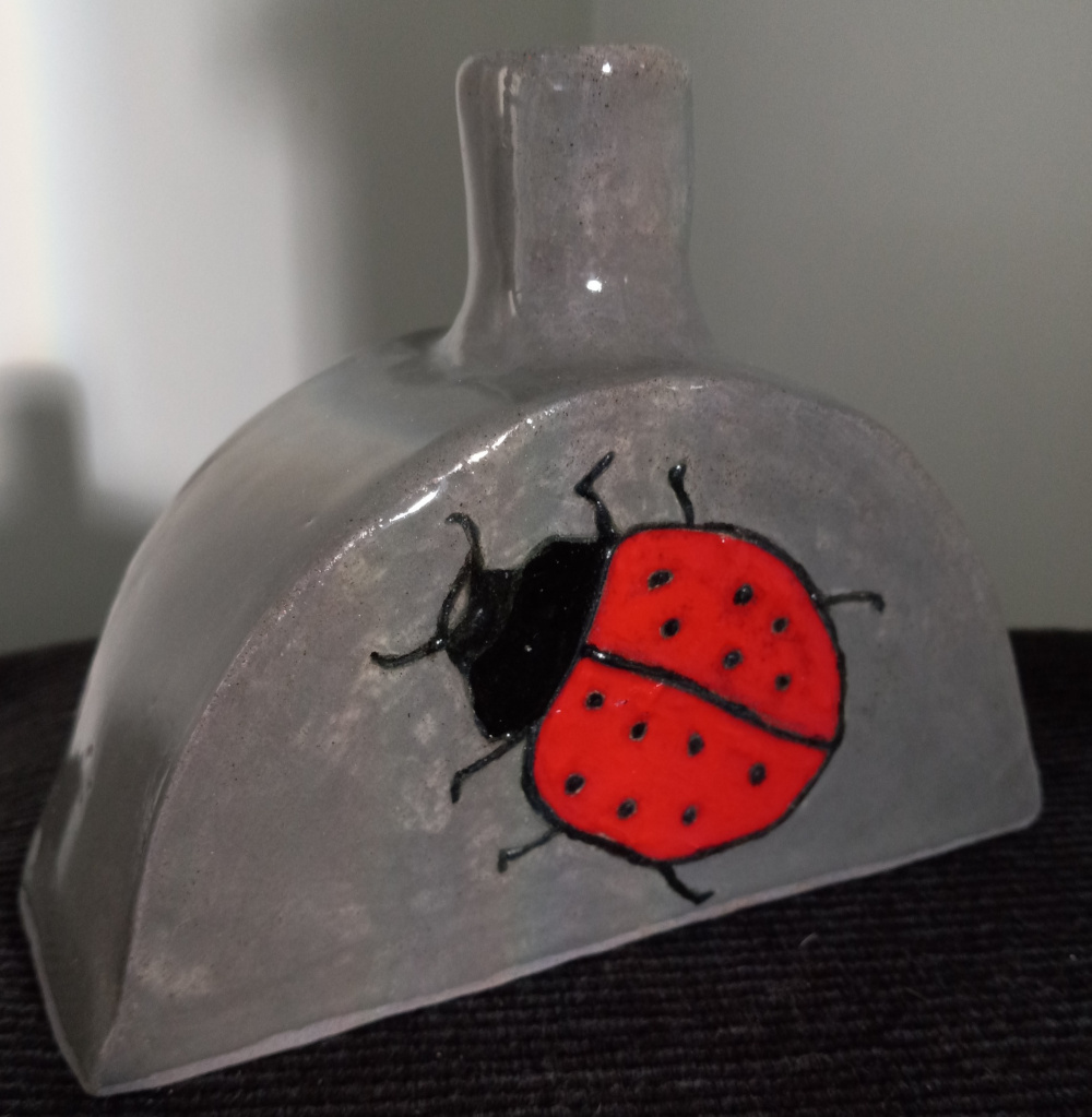 Vessel in granite clay with lady bug painted on using black slip and red underglaze.